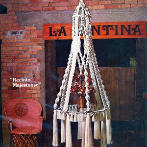 May include: A white macrame hanging planter with a wooden frame and a glass top. The planter is decorated with tassels and has a sign that reads "La Cantina" above it. The sign is red with black lettering. There is a sign below the planter that reads "Recinto Majestuoso".