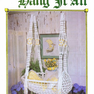 May include: A white macrame baby cradle hanging from the ceiling. The cradle is made of white rope and has a white wicker base. The cradle is decorated with yellow beads. The text "SNOW FLAKE CRADLE" is printed below the cradle.