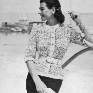 May include: A black and white photograph of a woman wearing a knitted cardigan with a white collar and buttons. She is also wearing a white belt with a large buckle and a bracelet. The cardigan has a speckled pattern.