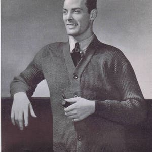 K&ouml;nnte beinhalten: Ein Schwarzwei&szlig;foto eines Mannes, der einen dunkelgrauen Strickpullover mit Kn&ouml;pfen und einer dunklen Krawatte tr&auml;gt. Er h&auml;lt eine Pfeife in seiner rechten Hand.