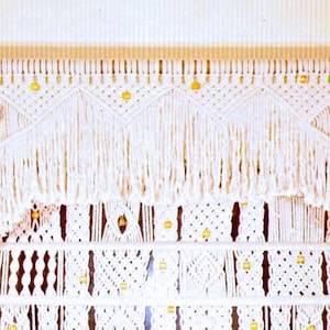 May include: White macrame wall hanging with a decorative fringe and gold beads. The macrame is made in a geometric pattern.