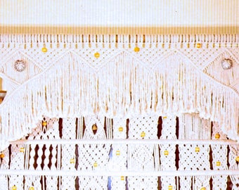 May include: White macrame wall hanging with a decorative fringe and gold beads. The macrame is made in a geometric pattern.