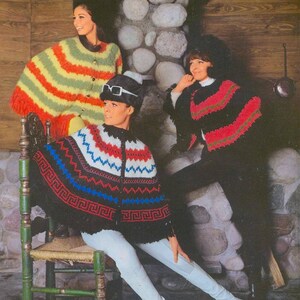 May include: Three women wearing colorful knitted ponchos. The woman in the center is sitting on a wooden chair. The woman on the left is wearing a striped poncho with yellow, orange, and green stripes. The woman on the right is wearing a black poncho with red, white, and blue stripes. All three women are wearing hats.