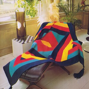 May include: A colourful geometric patterned crocheted throw blanket draped over a brown leather chair with wooden legs. The blanket features a large yellow diamond shape in the centre.
