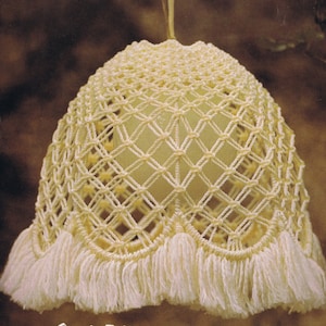 May include: A white macrame lampshade with a fringe trim. The lampshade is hanging from a white cord. The background is a blurry brown color.