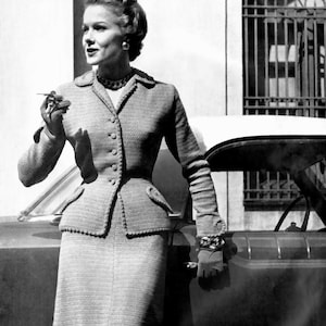 May include: A vintage black and white photograph of a woman in a tailored suit. The suit features a fitted jacket with button details and a matching pencil skirt. The woman is wearing a necklace and bracelet, and holding a cigarette.