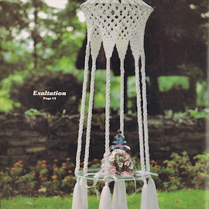 Può includere: Un portavasi sospeso in macramè bianco con un vaso di vetro rotondo e fiori rosa. Il portavasi ha un fiocco bianco su ciascuno dei sei cordini. Il titolo "Exaltation Page 14" è visibile nell'immagine.