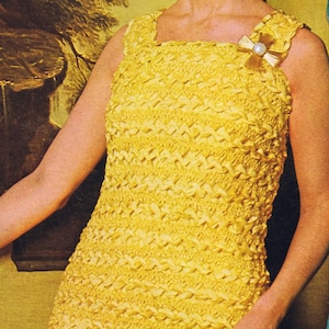 Può includere: Una donna indossa un abito di uncinetto giallo con una spilla dorata e bianca. L'abito ha una scollatura quadrata ed è senza maniche. La donna ha i capelli raccolti in uno chignon e indossa orecchini.