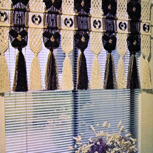 Puede incluir: Una cenefa de macramé para ventana con hilo de color marrón y crema, con un patrón geométrico repetitivo. La cenefa está colgada sobre un conjunto de persianas mini blancas.