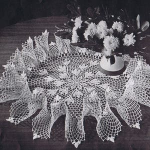 May include: A white crocheted doily with a scalloped edge and a delicate floral design in the center. The doily is laid out on a dark wooden surface.