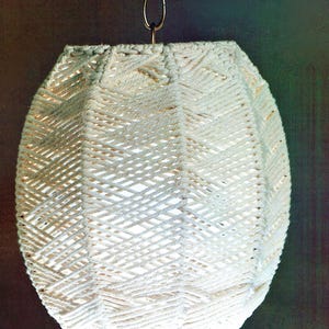 Op de afbeelding: Crèmekleurige macramé hanglamp. Het geweven ontwerp creëert een textuur, geometrisch patroon. Het licht hangt aan een metalen ketting. De vorm van de lamp is een langwerpig ovaal, geschikt voor een woonkamer of slaapkamer.