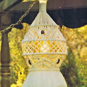 May include: A white macrame lampshade with a fringe bottom hangs from a gold chain. The lampshade is lit from within, casting a warm glow. The text "Moon Shadow Pattern on Page 6" is printed below the lampshade.