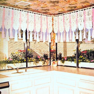 以下が含まれることがあります： 白いカウンタートップと白いキャビネットのある白いキッチン。キッチンには、ピンクのアクセントが付いた白いマクラメカーテンのある窓があります。窓の外には緑の植物のある庭があります。