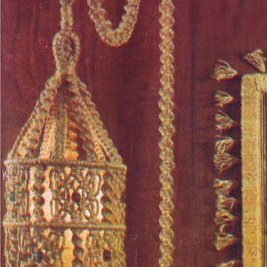 May include: A macrame hanging lampshade with a geometric design and fringe. The lampshade is made of natural fiber rope and hangs from a long rope with a tassel at the end. The lampshade is hanging on a dark red wall.