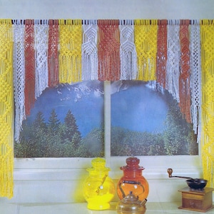 Pode incluir: Uma valance de macramé em amarelo, branco e vermelho pendura-se sobre uma janela com uma cena de montanha. Dois frascos de vidro, um amarelo e outro laranja, estão na janela, juntamente com um pequeno moedor de café e uma chaleira.