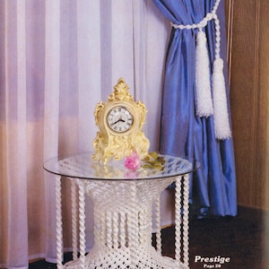 Può includere: Un tavolo in crochet bianco con un piano in vetro e un orologio dorato in cima. Il tavolo è davanti a una tenda blu con frange bianche. Il testo "Prestige Page 20" è visibile nell'angolo in basso a destra dell'immagine.