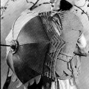 Puede incluir: Una fotografía en blanco y negro de una mujer con un sombrero y sosteniendo una sombrilla grande y redonda. La sombrilla está hecha de un material tejido y tiene un borla decorativa.