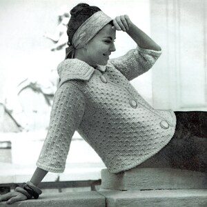 May include: A woman wearing a white knitted cardigan with a textured pattern and a large collar. She is wearing a headband and has her hand on her forehead. The cardigan has buttons down the front.