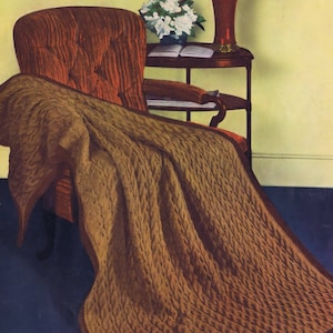 May include: A brown knitted throw blanket with a cable knit pattern draped over a brown armchair. The armchair is in front of a side table with a lamp and a bouquet of white flowers.