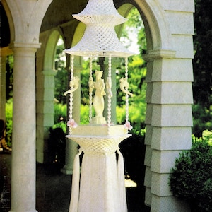 May include: A white macrame hanging sculpture with a fountain design. The sculpture features a white figure, pink flowers, and white tassels. The sculpture is hanging in a white archway with green foliage in the background. The title "Fountain of the Orient" is printed below the sculpture.