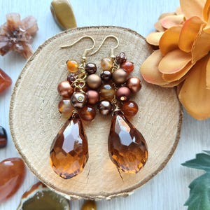 Pode incluir: Um par de brincos pendentes com cachos de contas castanhas, bronze e âmbar presos a ganchos de metal dourado. Cada brinco termina num grande pendente de vidro âmbar facetado em forma de lágrima que capta a luz.
