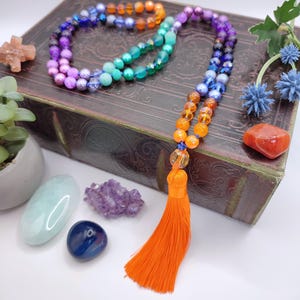 May include: A rainbow colored beaded necklace with an orange tassel. The necklace is made with different colored beads, including purple, blue, green, orange, and yellow. The necklace is lying on a brown book with a few crystals nearby.