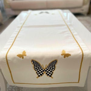 May include: Cream-coloured table runner with gold embroidered trim and butterfly designs. The central butterfly has a black and white chequered pattern, smaller butterflies are gold. The runner is on a white table.
