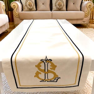 May include: Cream-colored table runner with black and gold trim. The runner features a gold embroidered monogram design. The runner is placed on a white table, with a sofa and pillows in the background. The runner is a decorative home accent.