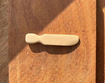 May include: A wooden butter knife with a rounded handle. The knife is light brown and has a smooth surface.