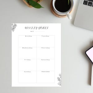 May include: A white weekly goals planner with floral accents. The planner is divided into sections for each day of the week, with the word "Prayers" at the bottom. A cup of coffee and a laptop are in the background.