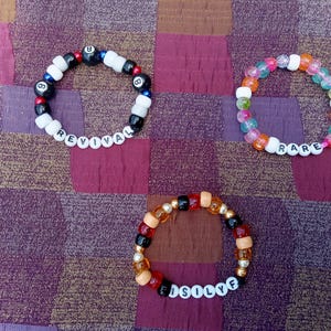 May include: Three beaded bracelets with different color schemes and text. One bracelet has black and white beads with the word "REVIVAL". Another has colorful beads with the word "RARE". The third has orange, black, and gold beads with the word "FISOLYF".