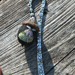 May include: A silver metal bookmark with a curved top and a decorative floral design. The bookmark features a small, black, oval-shaped charm with a painted scene of a house and flowers. A string of beads hangs from the top.