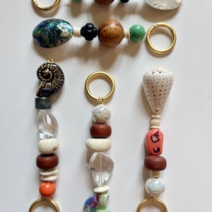May include: A collection of decorative keychains with various beads and charms. The keychains feature gold-colored metal rings, wooden beads, and unique charms like seashells and ceramic pieces. The color palette includes natural tones, greens, and pops of orange.
