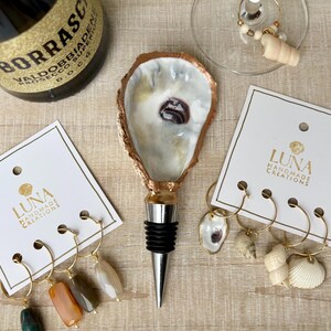 May include: A wine bottle stopper with an oyster shell design, a bottle of Borrasco Prosecco, and wine charms. The stopper has a silver base and a black rubber seal. The wine charms have gold hoops and shell and stone accents.