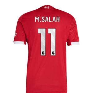 Może przedstawiać: Czerwona koszulka piłkarska z nazwiskiem "M. SALAH" i numerem "11" w kolorze białym. Koszulka ma białe wykończenia na rękawach i mały emblemat u góry. Koszulka jest w jednolitym czerwonym kolorze.