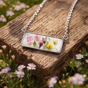 Peut inclure: Un collier argenté avec un pendentif rectangulaire orné de fleurs pressées roses, jaunes et rouges. Le pendentif est posé sur un fond blanc et repose sur une surface en bois, avec une chaîne visible.