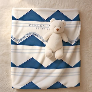 May include: A white fleece baby blanket with a blue chevron pattern and white stripes. The blanket has the text "Camden R. 11-24-20" and "Greater than the waves of trouble, love for you is standing." A white teddy bear is sitting on the blanket.