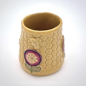 May include: A ceramic tumbler with a honeycomb pattern, bees, and a flower design. The tumbler is beige with purple and pink accents. The cylindrical cup is approximately 4 inches tall.