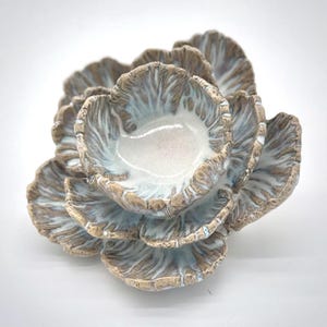 May include: A ceramic sculpture resembling a cluster of stylized flower petals. The petals are a blend of light brown and white, with a subtle blue-grey wash in the interior. The piece has a textured, organic appearance.