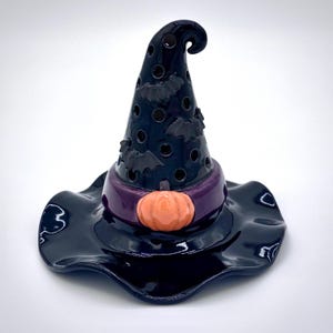 May include: A glossy black ceramic witch hat with a wide brim, purple band, and orange pumpkin accent. The hat features bat cutouts and is a decorative Halloween item.