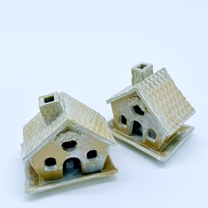 May include: Two ceramic miniature houses with tan roofs and walls, each with a chimney and windows. The houses sit on square bases. The roofs have a textured, tiled appearance, and the walls have a slightly weathered look.