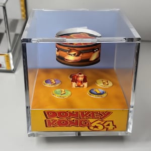 以下が含まれることがあります： ドンキーコング64をテーマにした透明なアクリル製キューブディスプレイケース。中にはドンキーコングのミニチュアフィギュアが番号付きトークンのある黄色い台座の上に立ち、上部には樽のグラフィックが吊り下げられています。台座には「DONKEY KONG 64」の文字が記されています。