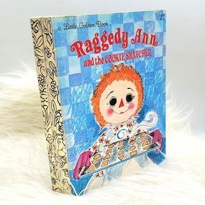 May include: A Little Golden Book titled "Raggedy Ann and the Cookie Snatcher" featuring a red-haired Raggedy Ann doll holding a baking sheet of cookies.
