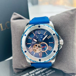 May include: A Bulova Marine Star automatic watch with a blue dial, silver bezel, and blue rubber strap. The watch features Roman numeral hour markers, a visible movement, and the text "BULOVA Marine Star".