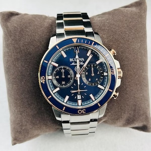 May include: A Bulova Marine Star watch with a stainless steel band and a blue dial. The watch features gold-tone accents on the bezel and chronograph dials. The watch is displayed on a brown pillow.