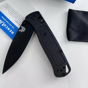 May include: A black pocket knife with a black handle and a partially open blade. The knife has a textured handle and a small butterfly logo on the blade. The knife is on a white surface.