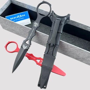 Könnte beinhalten: Zwei schwarze taktische Messer mit schwarzen Scheiden. Das Messer links hat einen roten Griff und ein Schmetterlingsdesign. Das Messer rechts hat einen schwarzen Griff und den Text "BENCHMADE" an der Seite. Beide Messer befinden sich in einer schwarzen Box mit einem weißen Etikett, auf dem "BENCHMADE USE AND CARE MANUAL" steht.