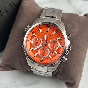Op de afbeelding: Zilverkleurig Bulova horloge met een oranje wijzerplaat en een roestvrijstalen band. Het horloge heeft meerdere subwijzerplaten en witte markeringen. Het horloge wordt gepresenteerd op een bruin kussen.