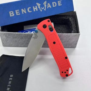 Puede incluir: Un cuchillo plegable Benchmade rojo con una hoja plateada y un clip de bolsillo negro. El cuchillo está abierto y la hoja está parcialmente extendida. El cuchillo está sobre un paño negro con la palabra "Benchmade" impresa en él.