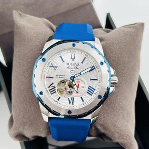 May include: A Bulova Marine Star automatic watch with a silver-tone stainless steel case and a blue silicone strap. The watch features a white dial with Roman numeral hour markers and a visible movement. The watch is water-resistant to 200m.